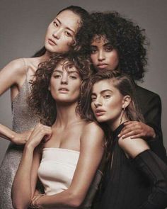 three women are posing together in front of a gray background and one woman has her arms around the other