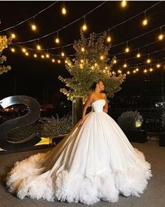 a woman in a white wedding dress standing on a brick walkway with lights strung above her