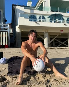 a shirtless man sitting on the beach holding a white ball in front of a large building