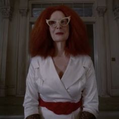 a woman with red hair and glasses standing in front of a door wearing a white suit