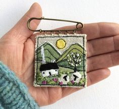 a hand holding a small square brooch with two cows on it and mountains in the background