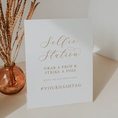 a white card with gold lettering on it next to a vase filled with dry grass