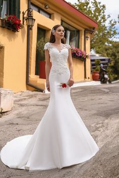 a woman standing in front of a yellow building wearing a white wedding dress with flowers on it