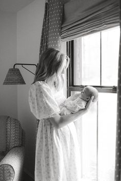 a woman holding a baby standing next to a window