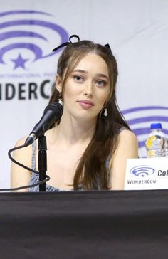 a woman sitting at a table with a microphone in front of her and a sign behind her