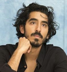 a man with long curly hair and a black shirt is looking at the camera while he has his hand on his chin