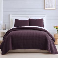 a bed with purple bedspread and pillows in a white room next to a night stand