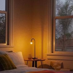 a bedroom with a bed, table and lamp in the corner by two large windows