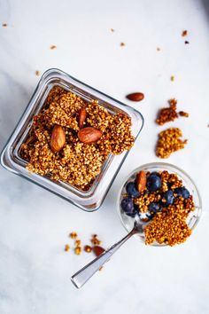 granola with nuts and blueberries in a glass dish next to a spoon on a white surface