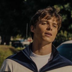 a young man standing in front of a car looking off into the distance with trees and grass behind him