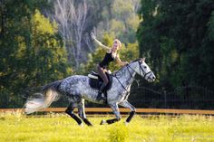 a woman riding on the back of a white horse through a lush green field with trees in the background