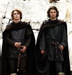 two men standing next to each other in front of a stone wall wearing medieval clothing