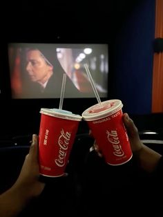 two red cups with straws are held in front of a television screen and the person is watching