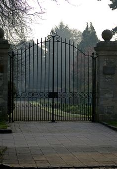 an iron gate is open on the side of a brick walkway with trees in the background