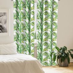 a white bed sitting next to a window covered in green leafy curtain panels on top of a wooden floor