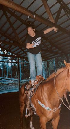 a woman standing on top of a brown horse in a barn next to another horse