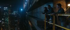 three men are standing on the edge of a high rise building at night, looking out over the city