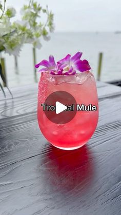 a pink drink sitting on top of a wooden table next to the ocean with purple flowers