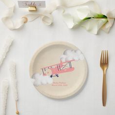 a plate with a pink plane on it next to some white flowers and gold utensils