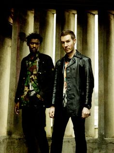 two men standing next to each other in front of columns and pillars with one man wearing a leather jacket