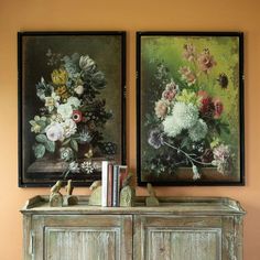 two paintings are hanging on the wall next to a wooden cabinet with books and vases