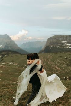 a bride and groom hugging on top of a mountain with their veil blowing in the wind