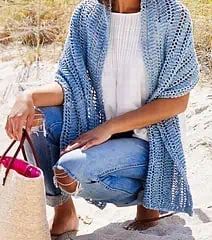 a woman sitting in the sand with a bag and purse on her lap, wearing a blue knitted cardigan