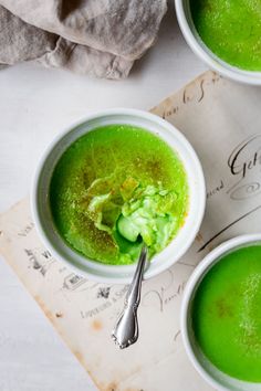 three white bowls filled with green soup on top of a paper towel next to two spoons