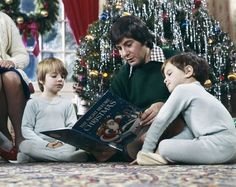 a man sitting on the floor with two children reading a book in front of a christmas tree