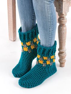 a woman standing next to a wooden table wearing green knitted boots with yellow flowers on them