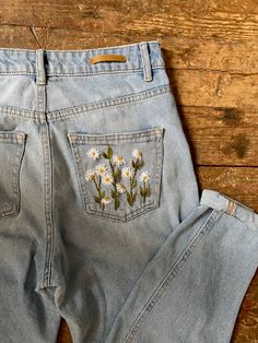 a pair of jeans with daisies embroidered on the front and back pockets, sitting on a wooden floor