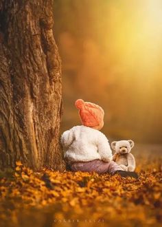 two teddy bears sitting next to a tree in the fall with leaves on the ground