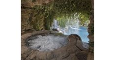 an outdoor hot tub in the middle of a pool surrounded by rocks and trees with water cascading over it