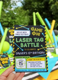 a person holding up a game on laser tag birthday card in front of some balloons