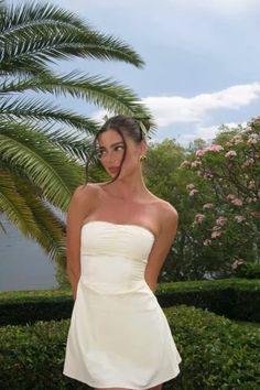 a woman in a short white dress standing next to a palm tree and shrubbery