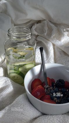 a white bowl filled with fruit next to a jar of pickles and cucumbers