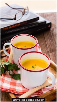 two white mugs filled with soup sitting on top of a wooden tray next to a stack of books