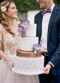 a bride and groom holding a wedding cake with purple flowers on top, in front of them