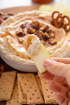 a hand holding an apple slice with pretzels and dip in the background