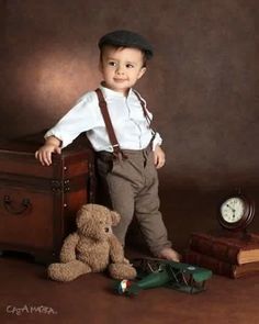 a little boy standing next to a teddy bear in front of an old suitcase and clock