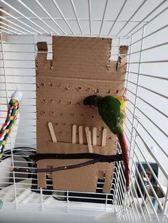 a green and yellow parakeet sitting on top of a cardboard box in a cage