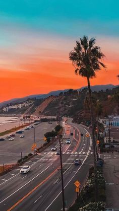 the sun is setting on an ocean side road with palm trees and cars driving down it