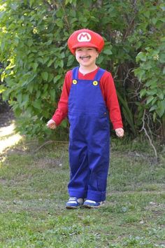 a young boy wearing a mario hat and overalls in front of some green bushes