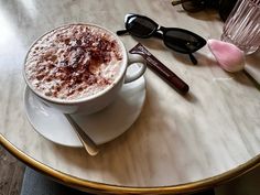 a cappuccino sits on a saucer next to sunglasses and a pink rose