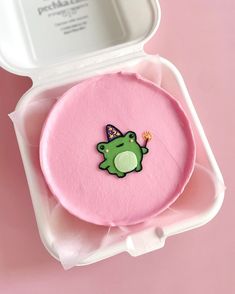 a pink cake with a green frog on it in a white box next to a mirror