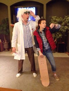 two people standing in a living room with a skateboard on the floor and one person wearing a white wig