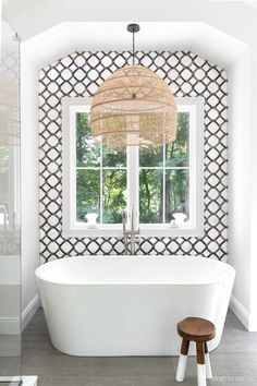 a white bath tub sitting under a window next to a wooden stool and lamp fixture