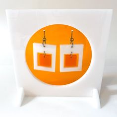 two square shaped orange and white earrings with silver earwires hanging from hooks on an acrylic stand