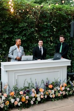 three men in suits are sitting at a white reception table with laptops on it