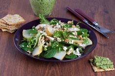 a purple plate topped with salad next to crackers and a cup of pesto